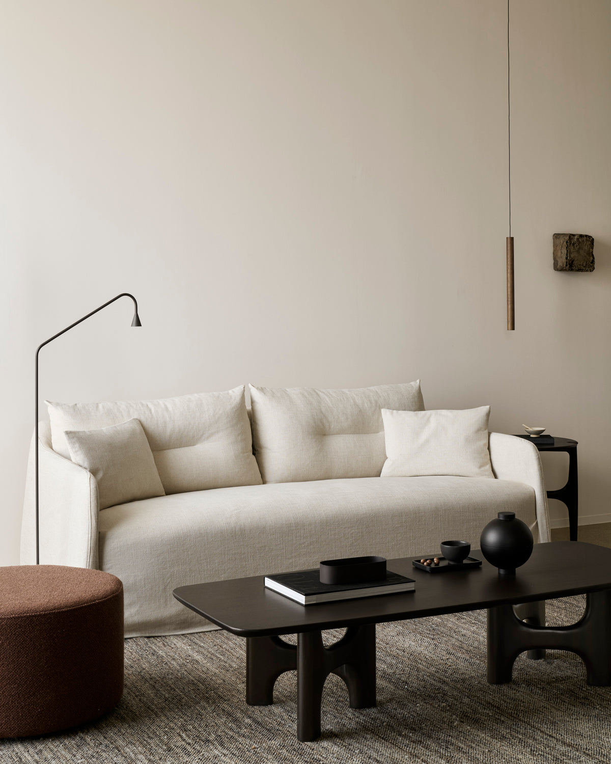 Minimalist living room with the Ethnicraft Weave sofa | 2 Seater Bone featuring linen-blend upholstery and removable covers, a black coffee table, brown round ottoman, black floor lamp, and neutral decor on a textured rug against a beige wall.