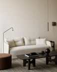 Minimalist living room with the Ethnicraft Weave sofa | 2 Seater Bone featuring linen-blend upholstery and removable covers, a black coffee table, brown round ottoman, black floor lamp, and neutral decor on a textured rug against a beige wall.
