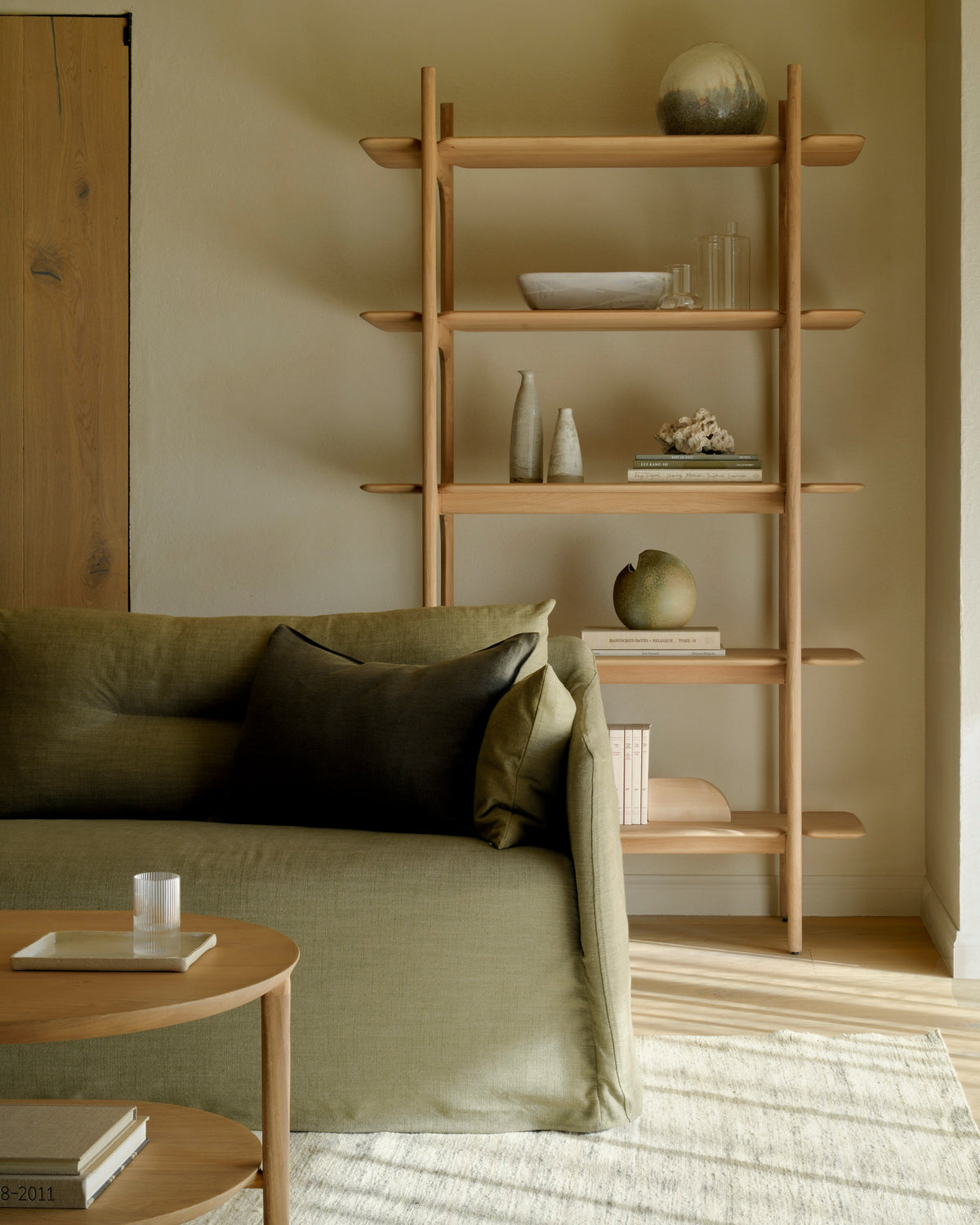 A minimalist living room features the Ethnicraft Weave Sofa | 2 Seater Green with removable covers, paired with a round wooden coffee table and a wooden shelf displaying decorative ceramics, books, and vases against a neutral wall. Warm natural light fills the cozy, modern space.