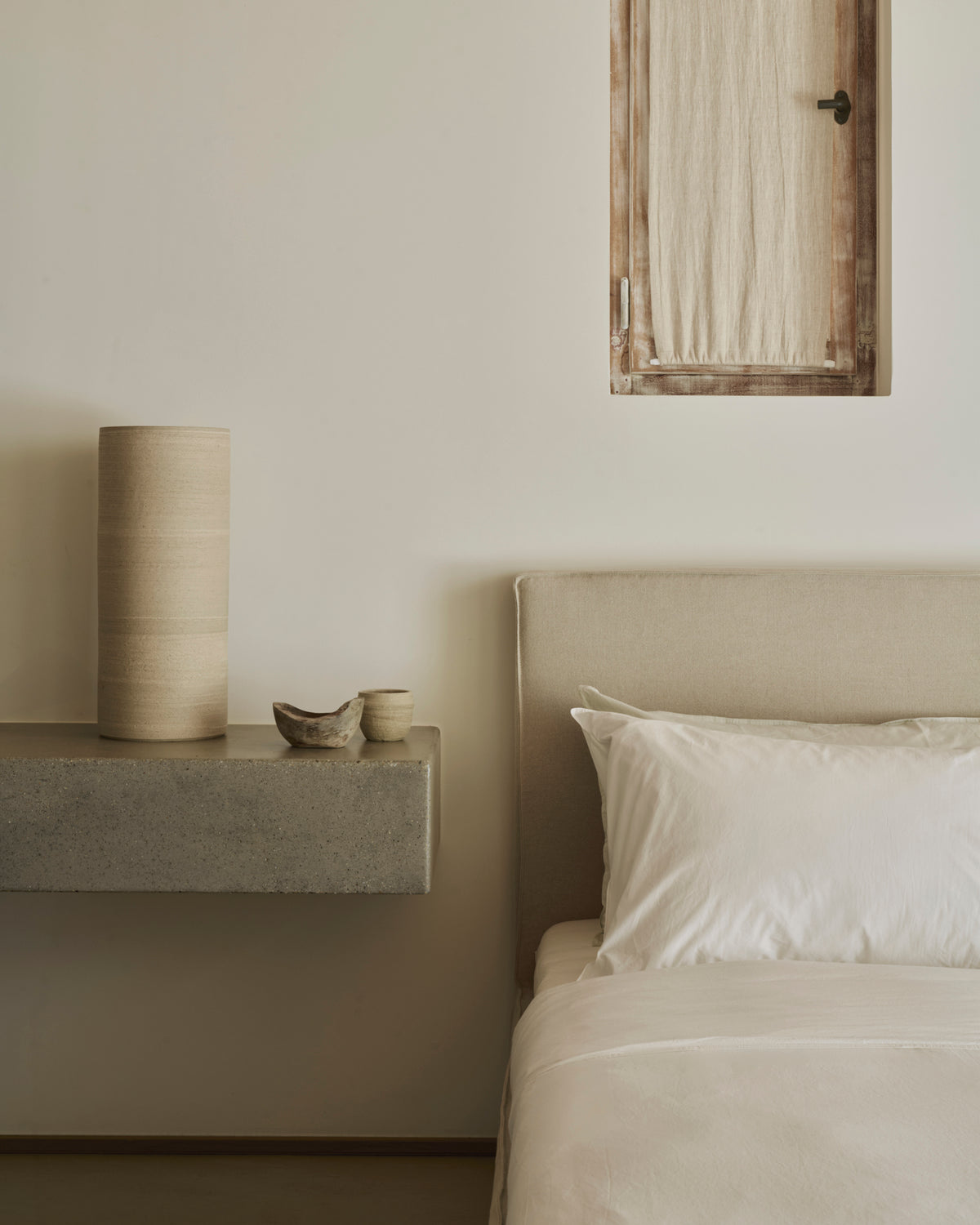 Minimalist bedroom featuring the Ethnicraft Revive Bed - Sand, outfitted with 100% linen removable covers, complemented by crisp white bedding, a concrete shelf displaying neutral-toned pottery, and a small window dressed with a beige curtain set against a light-colored wall.