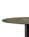 A close-up of the Ethnicraft Blend Dining Table - Ray Whisky shows its round, textured wooden top with visible grain and a dark cylindrical metal leg—ideal for commercial settings, set against a white background.
