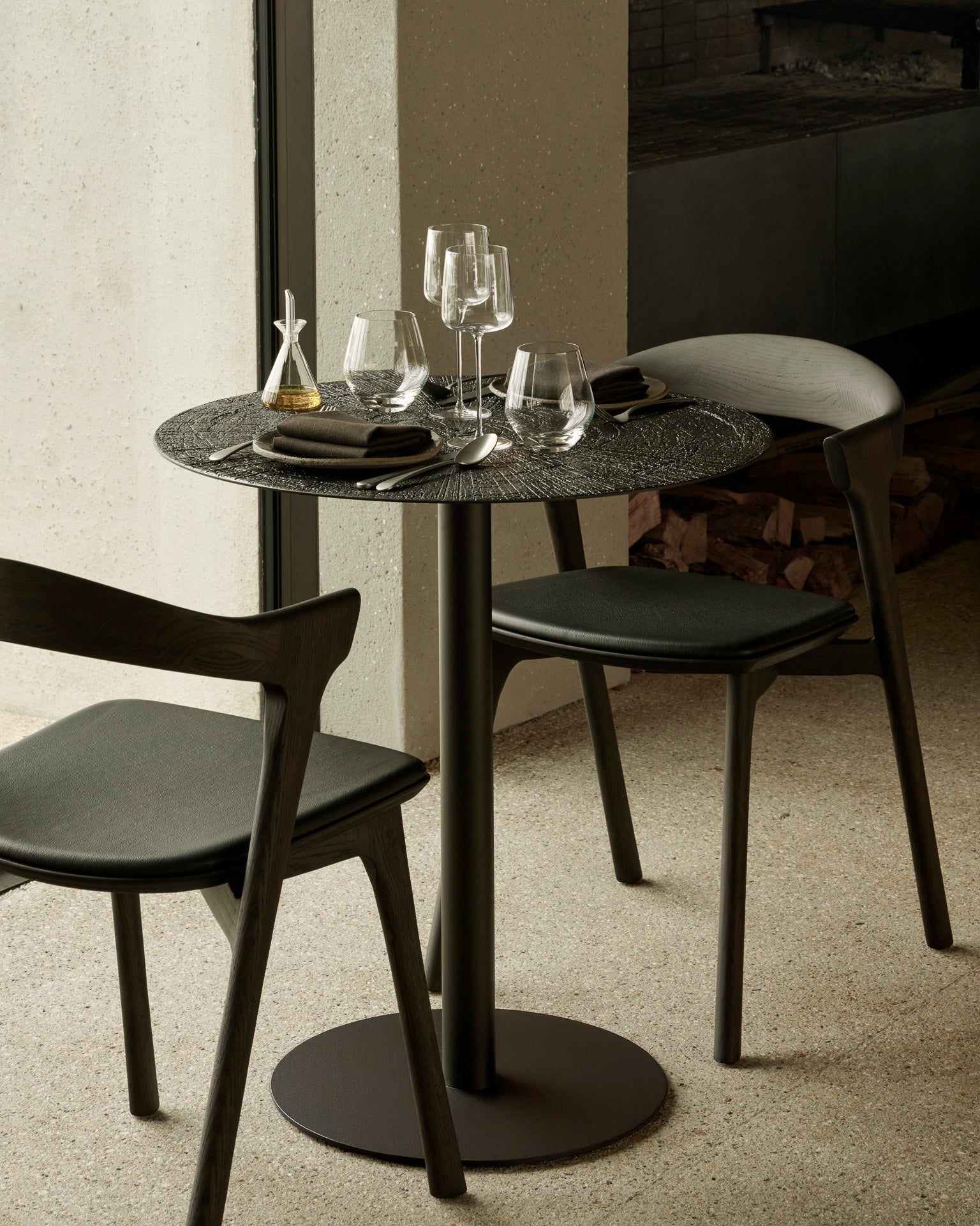 The Ethnicraft Blend Dining Table - Ray Whisky seats two and features modern wooden chairs, interchangeable table tops, dark plates, wine glasses, and linen napkins. Ideal for commercial spaces with soft lighting near a window.