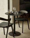 The Ethnicraft Blend Dining Table - Ray Whisky seats two and features modern wooden chairs, interchangeable table tops, dark plates, wine glasses, and linen napkins. Ideal for commercial spaces with soft lighting near a window.