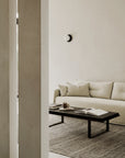 A minimalist living room with beige walls features the Ethnicraft Weave Sofa in Bone linen-blend fabric, a dark wooden coffee table topped with books and decor, a gray rug, and a round wall sconce for a calm, uncluttered vibe.