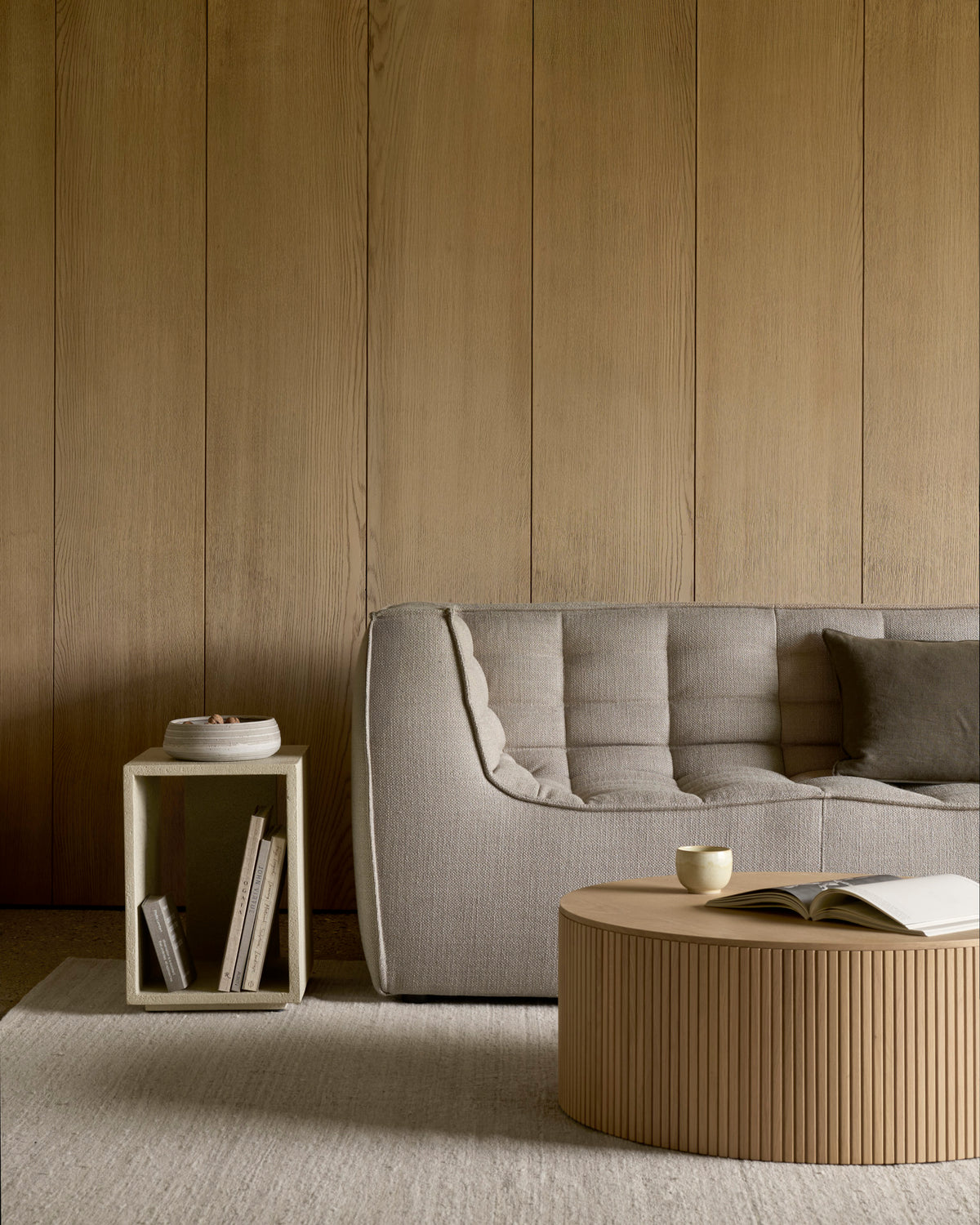 A modern living room features the N701 2-Seater Sofa in Beige by Ethnicraft, celebrated for its European craftsmanship and modular design. The space also includes a small side table topped with books and a bowl, alongside a round coffee table displaying an open book and a cup. Light wood paneling and neutral tones complete the serene atmosphere.
