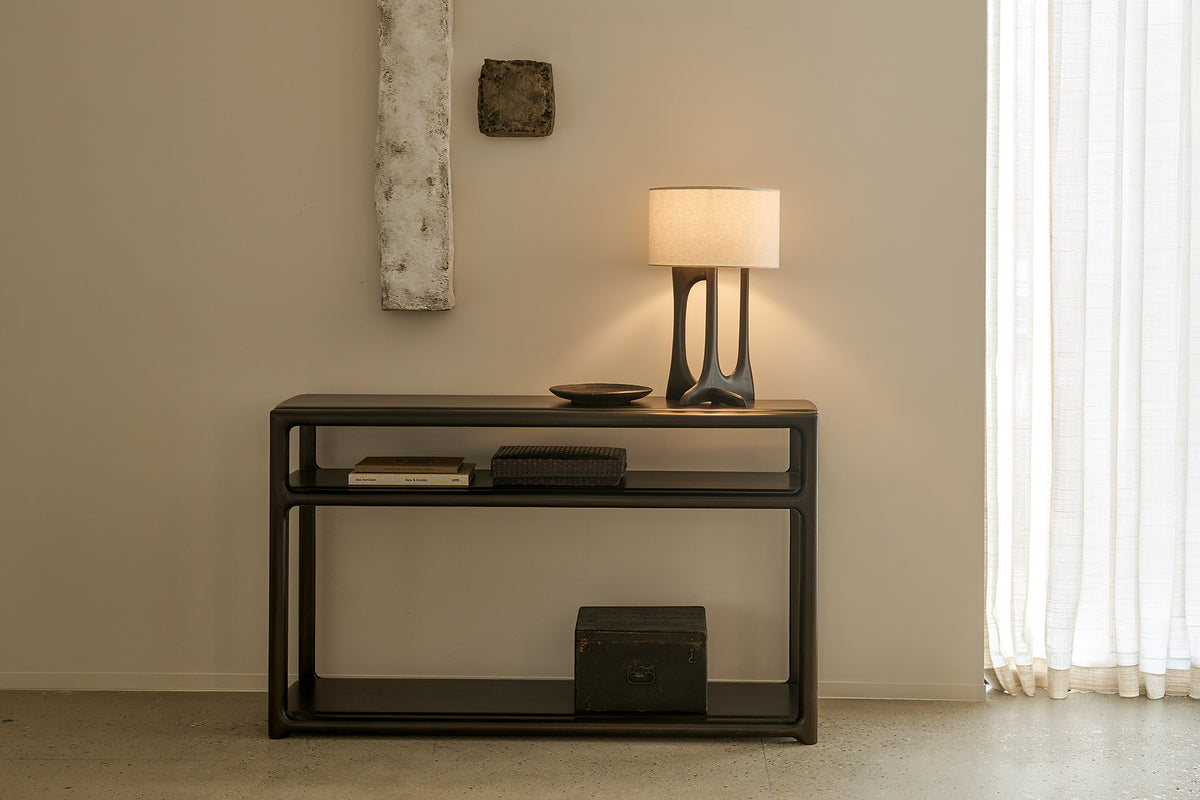 Dark mahogany Eda Console with two tiers, topped with a sculptural lamp, a small bowl, and books, with a storage box on the lower shelf.