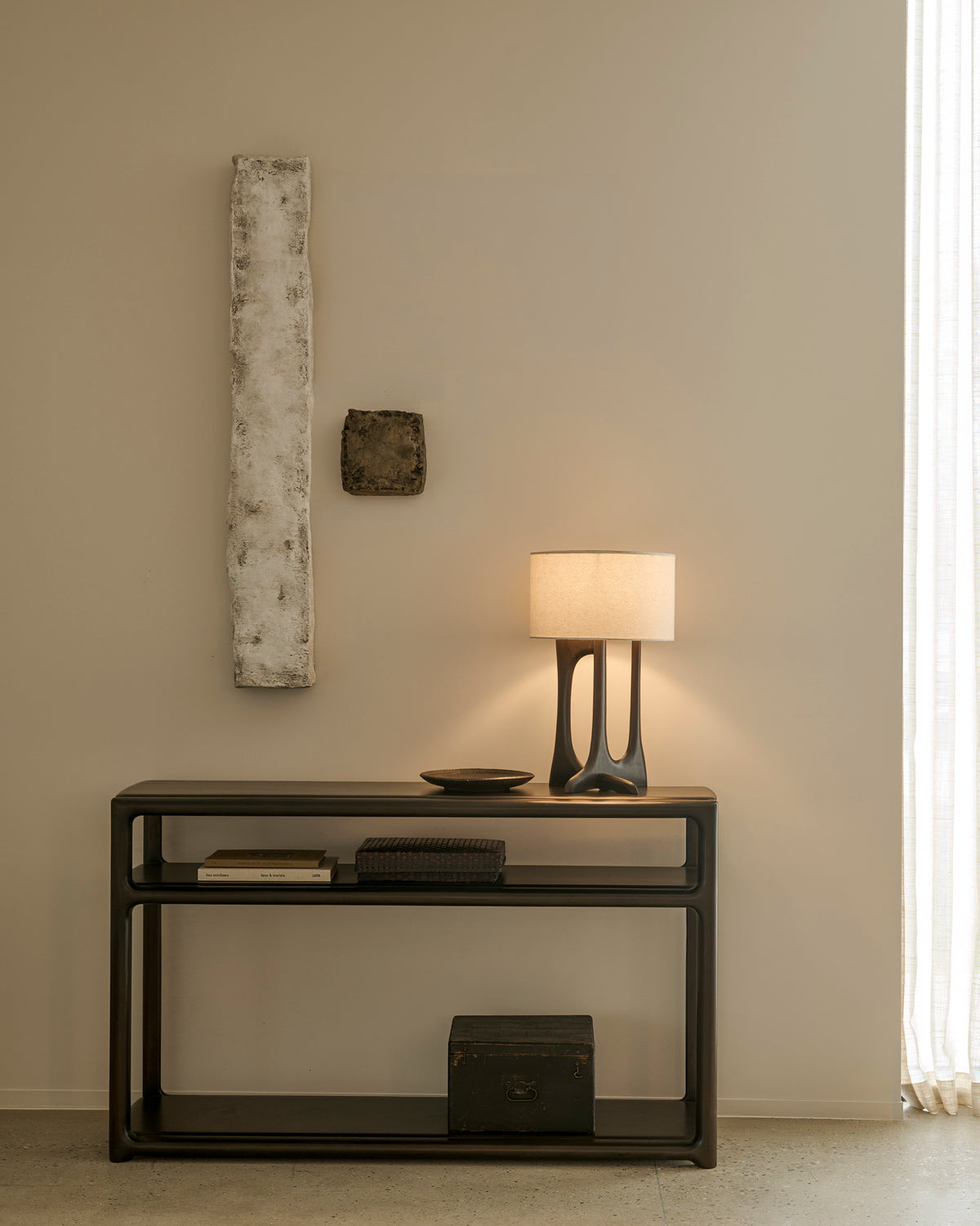 An heirloom-quality Ethnicraft Eda Console table with an open middle shelf, holding a lamp, a small bowl, a book, and two boxes, positioned beneath two wall-mounted sculptures.