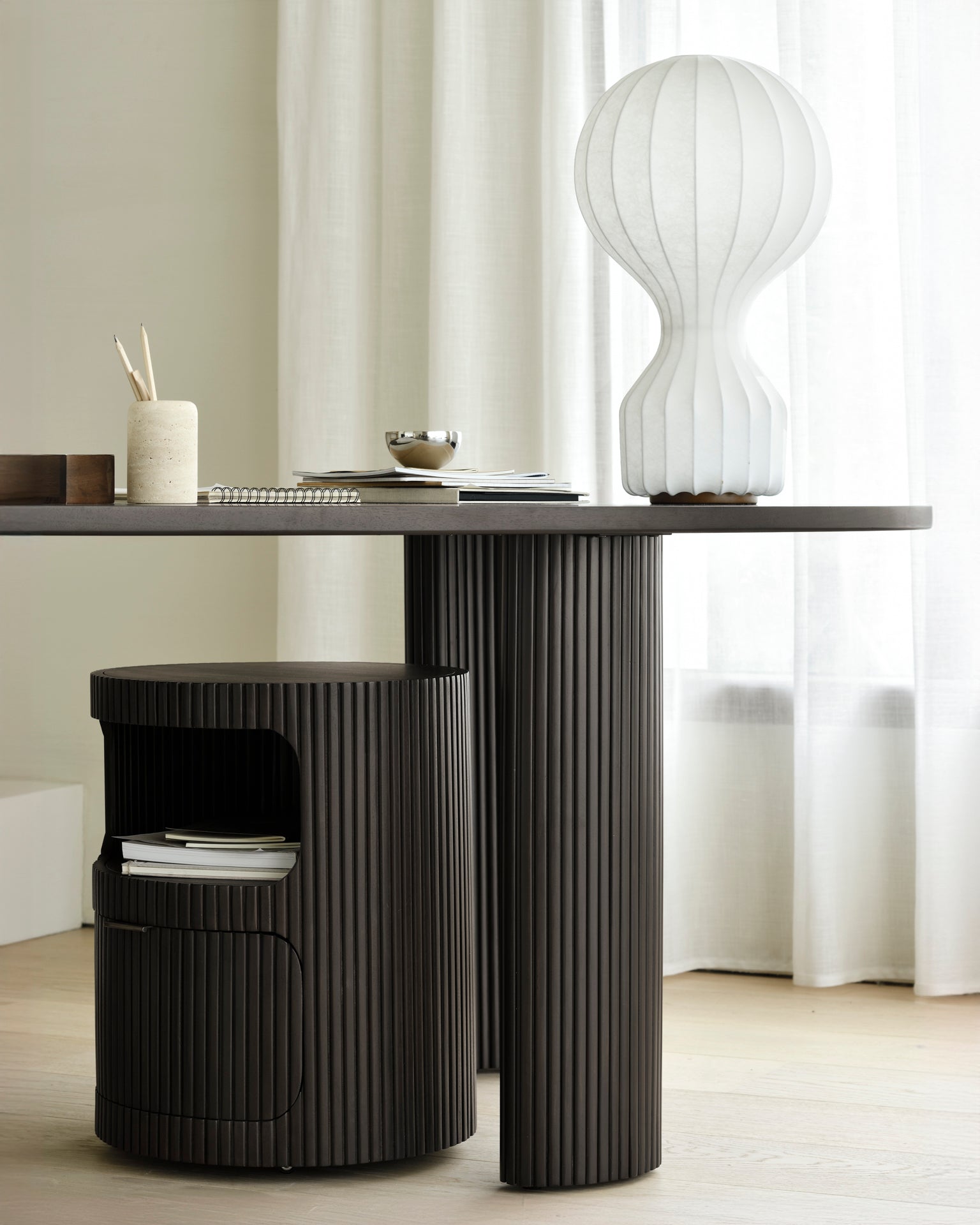 A modern dark brown Boomerang Drawer Unit by Ethnicraft with vertical grooves stands on a light wood floor, while a matching grooved storage stool sits beneath, softly framed by white curtains in the background.