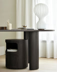 A modern dark brown Boomerang Drawer Unit by Ethnicraft with vertical grooves stands on a light wood floor, while a matching grooved storage stool sits beneath, softly framed by white curtains in the background.