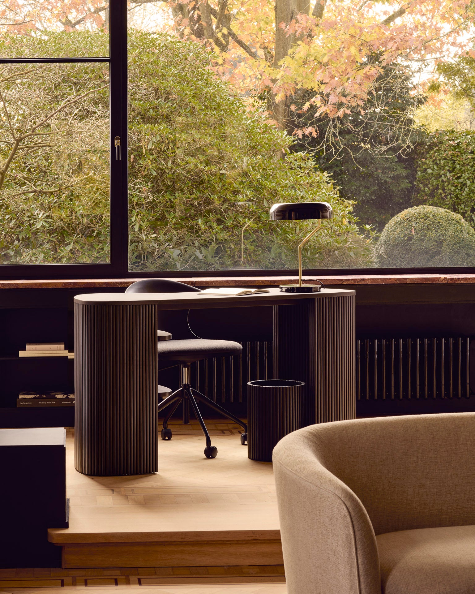 A modern home office features the Ethnicraft Roller Max Desk, an office chair, a black desk lamp, and a large window revealing lush greenery and autumn leaves. A beige armchair sits in the foreground.