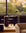 Modern home office featuring stylish furniture—a sleek brown mahogany Ethnicraft Roller Max Desk, paired with a lamp and chair—positioned by a large window overlooking lush greenery and autumn trees. A beige armchair is partially visible in the foreground.