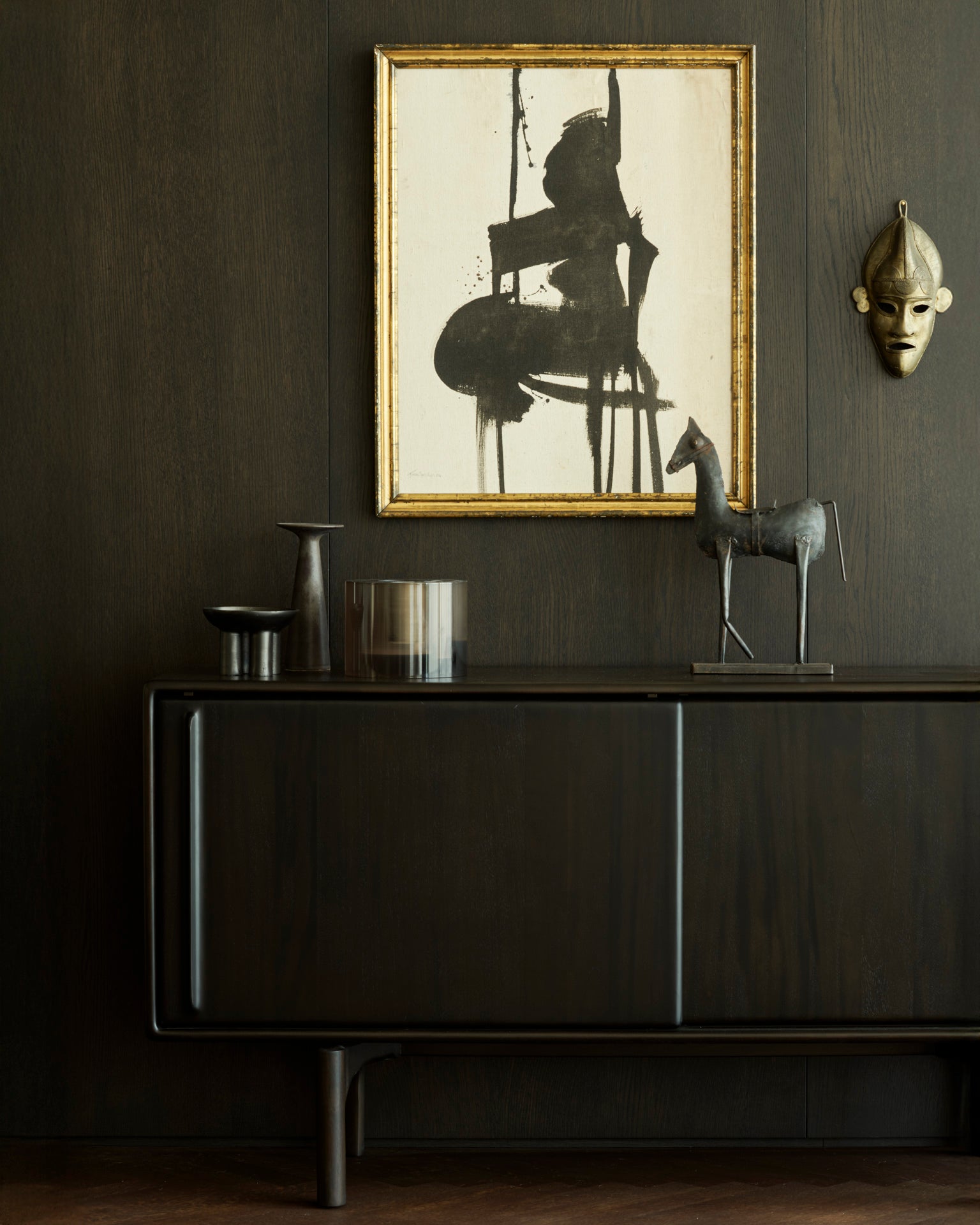 The Flow Sideboard by Ethnicraft stands against a matching wall, displaying decor like vases, a glass, and a metal horse statue. Above it, an abstract gold-framed painting and mask complete the vintage-modern look.