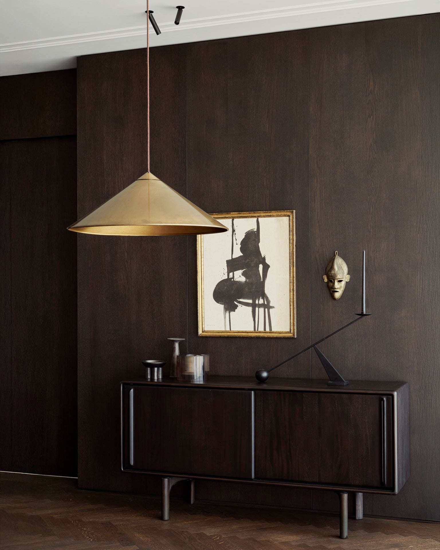 A modern interior features dark wood walls, a brass pendant lamp, and the Flow Sideboard by Ethnicraft displaying vases and minimalist sculpture. Above it, abstract art in a gold frame and a decorative mask adorn the wall.