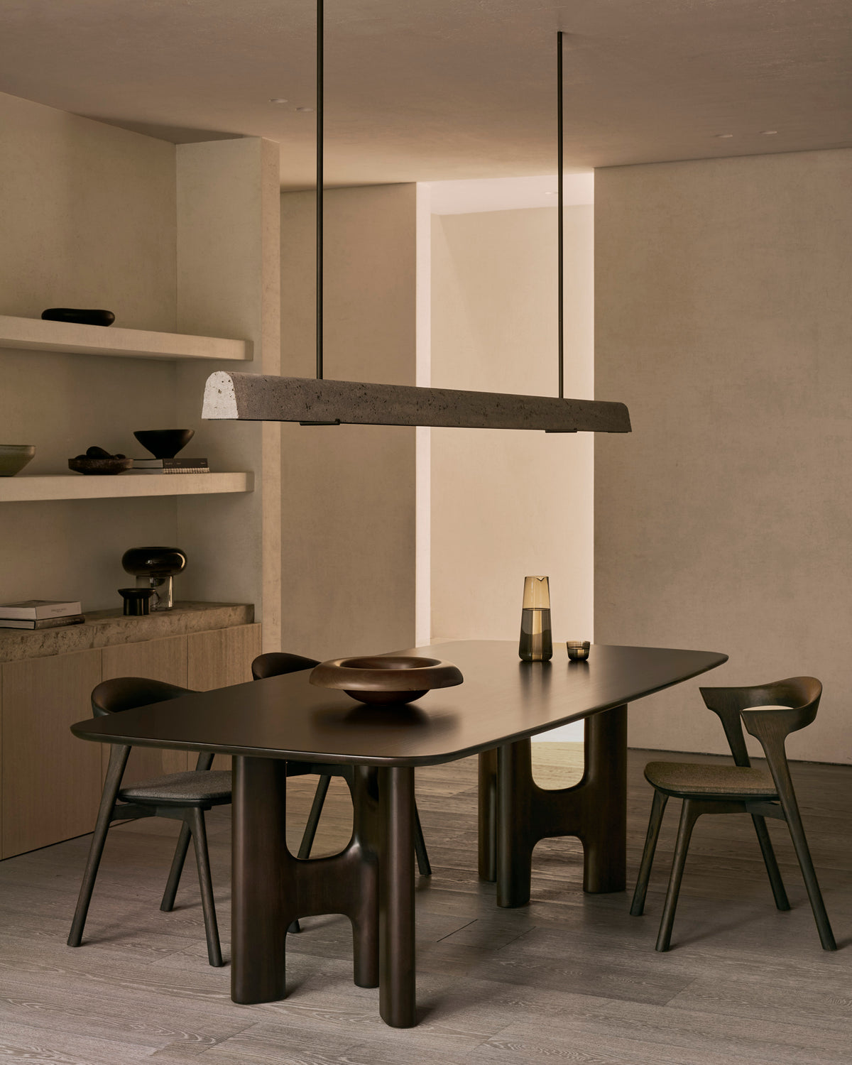 An Ethnicraft Flow Dining Table | Mahogany Rose Brown with rounded corners, surrounded by four wooden chairs in a minimalist room beneath a long, horizontal pendant light.