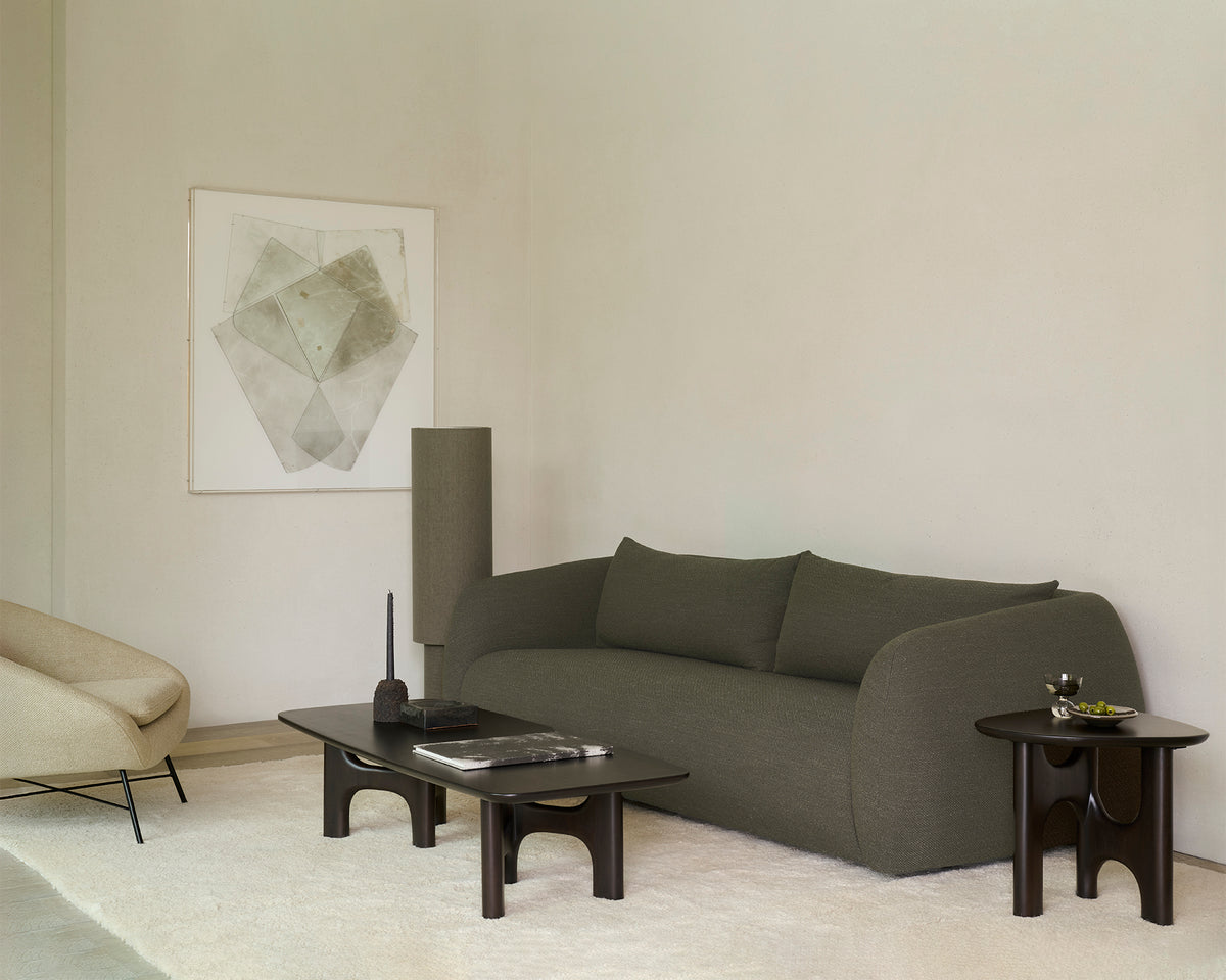 Minimalist living room featuring the Ethnicraft Figur Sofa | 3 Seater in green, abstract artwork on the wall, a beige armchair, and dark wood tables atop a light rug. Décor accents include a tray, candle, small sculptural object, and a plate with green fruit—perfect for contemporary living.