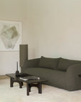 Minimalist living room featuring the Ethnicraft Figur Sofa | 3 Seater in green, abstract artwork on the wall, a beige armchair, and dark wood tables atop a light rug. Décor accents include a tray, candle, small sculptural object, and a plate with green fruit—perfect for contemporary living.