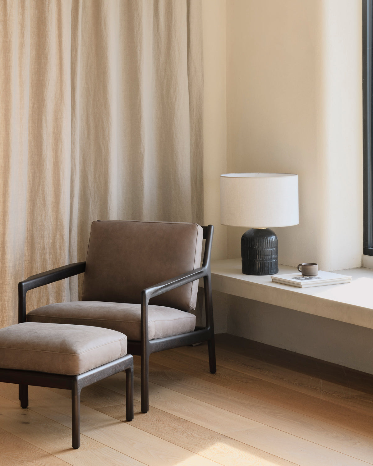The Jack Lounge Chair - Nubuck Leather by Ethnicraft sits on wooden flooring next to a white table, showcasing Scandinavian modernism against a beige curtain and cream wall.