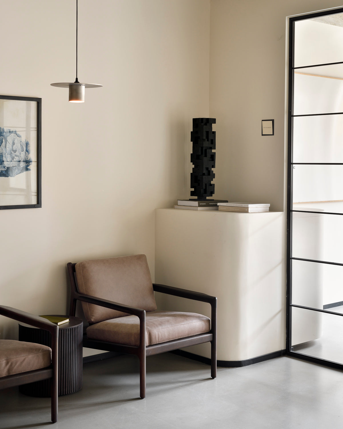 Minimalist room with two Ethnicraft Jack Lounge Chair - Nubuck Leather, a small side table, framed wall art, sculpture on a white pedestal, and glass door—decor reflecting Scandinavian modernism in neutral tones.