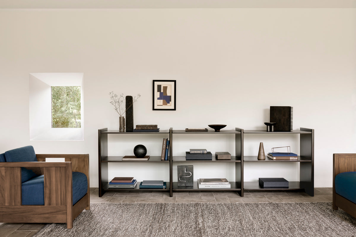 Minimalist living room featuring wooden shelves displaying books, vases, and decor accents, two Ethnicraft Roan Lounge Chairs in Ink with blue cushions inspired by vintage design, a gray rug, and a small window on a plain white wall that allows natural light to fill the space.