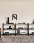 Minimalist living room featuring wooden shelves displaying books, vases, and decor accents, two Ethnicraft Roan Lounge Chairs in Ink with blue cushions inspired by vintage design, a gray rug, and a small window on a plain white wall that allows natural light to fill the space.