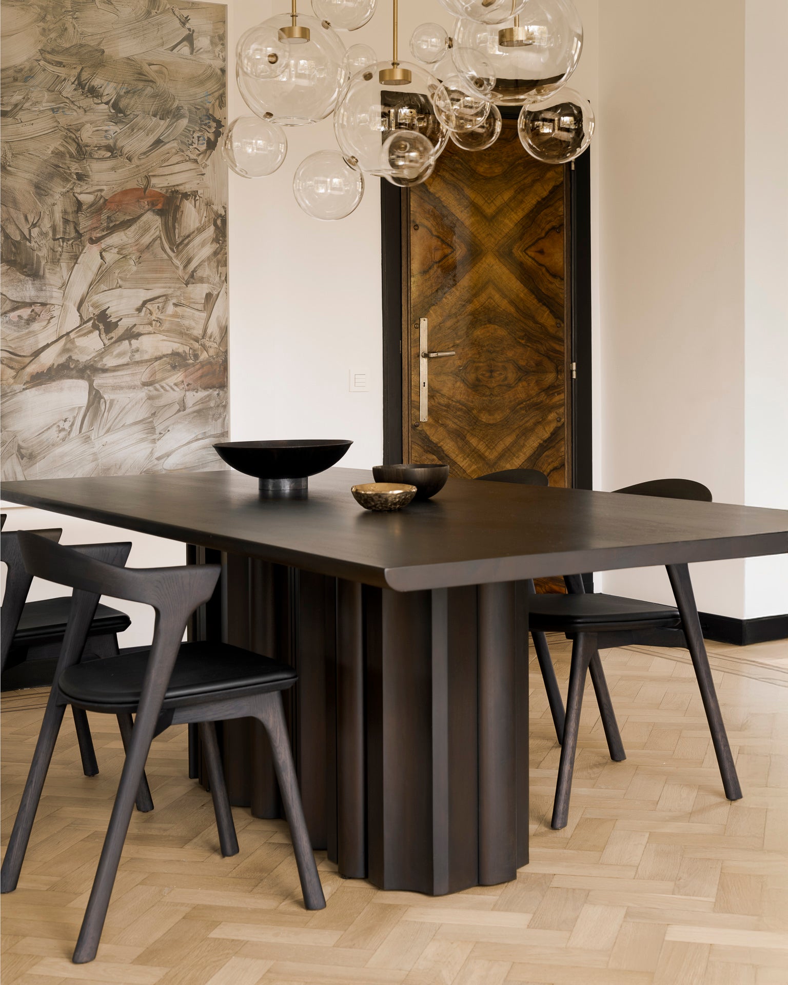 A modern dining area with Ethnicraft's Sequence Dining Table, matching chairs, glass globe pendants, abstract wall art, a geometric wooden door, and a light herringbone floor.