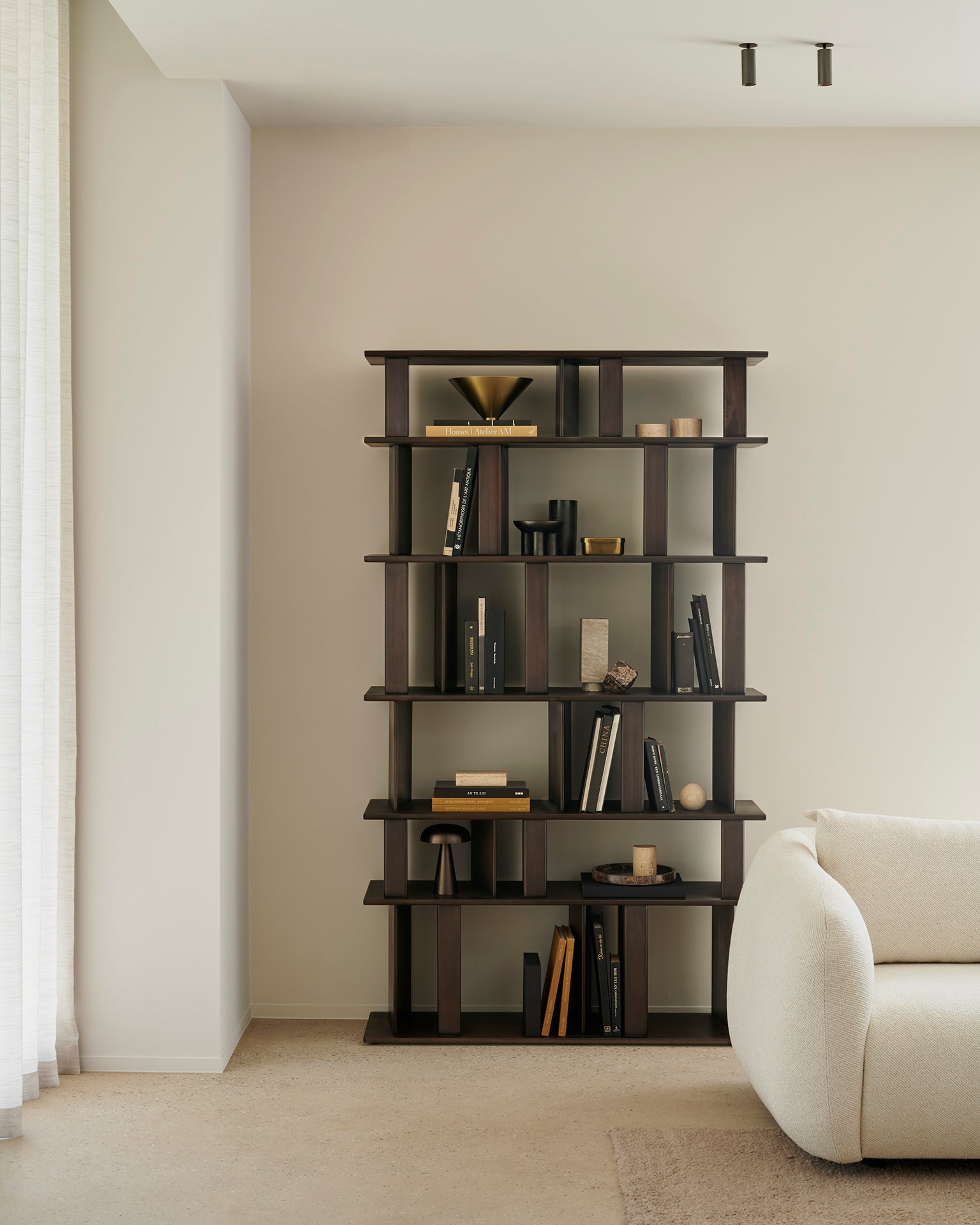 A modern living room features the tall Block Rack bookshelf by Ethnicraft in solid mahogany, displaying books and decor. A cream sofa is partially seen on the right, with sunlight entering through sheer curtains on the left.