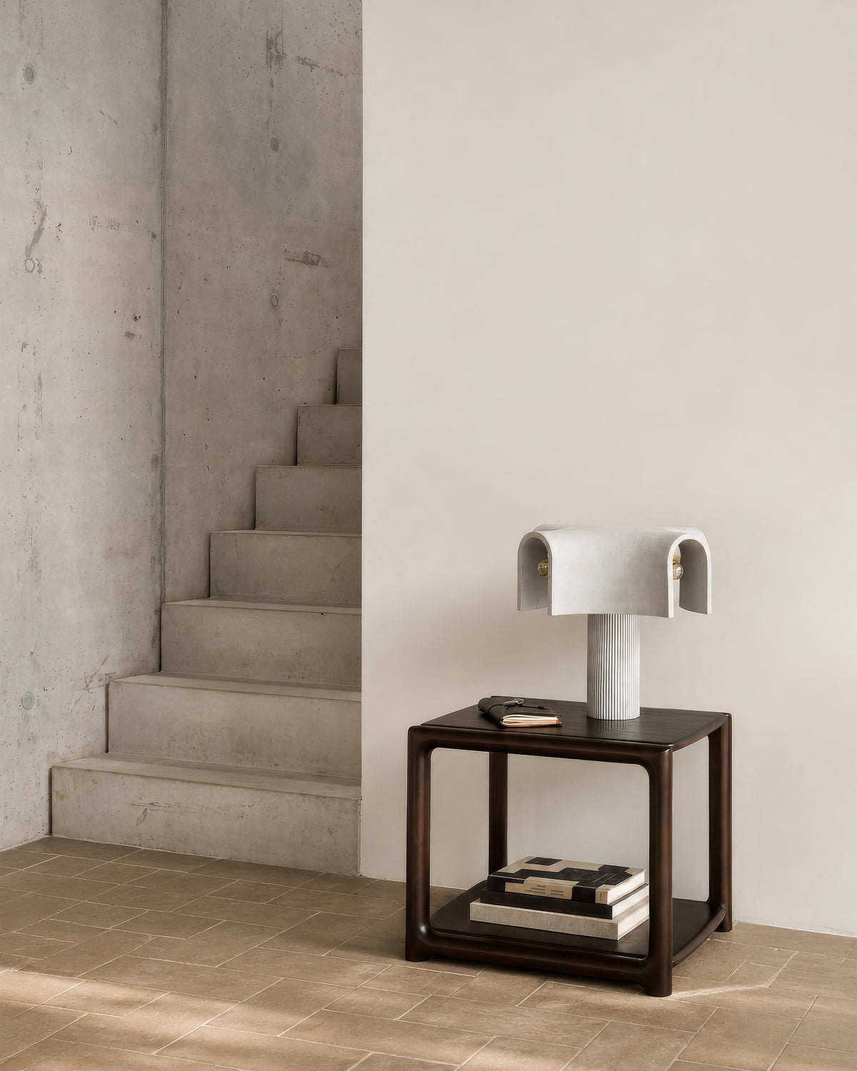 Dark mahogany Eda Side Table by Ethnicraft with a fluted pillar lamp and books on the lower shelf, positioned beside concrete stairs.