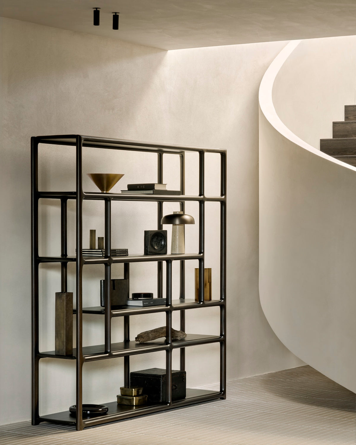 A tall, open-frame Ethnicraft Eda rack styled with books, small sculptures, and bowls, positioned beside a curved staircase.