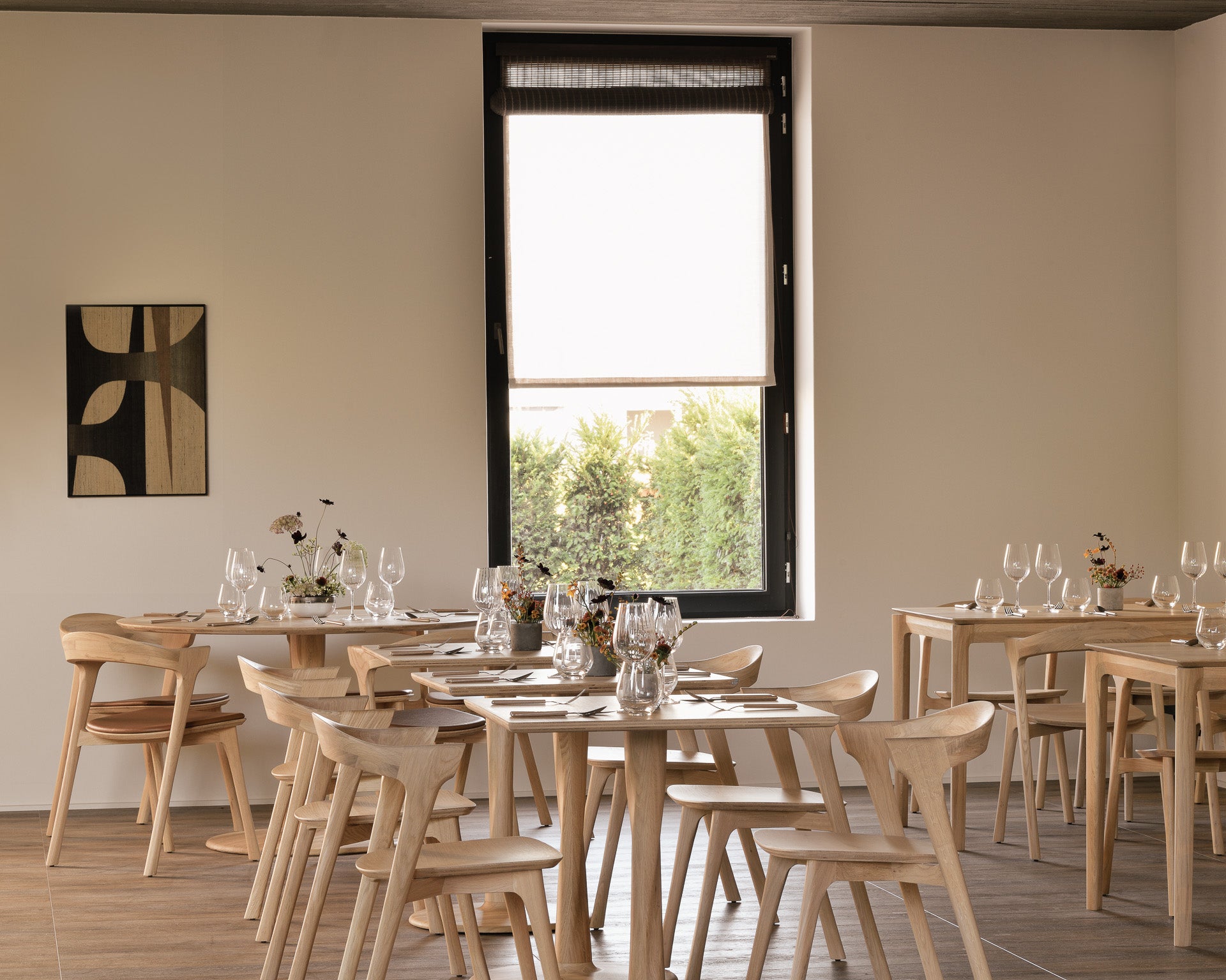 A minimalist restaurant features light wooden chairs and glassware atop the Ethnicraft Bok Dining Table in varnished oak. A large window lets in natural light, while abstract art and small flower vases adorn the beige wall.