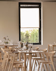 A minimalist restaurant features light wooden chairs and glassware atop the Ethnicraft Bok Dining Table in varnished oak. A large window lets in natural light, while abstract art and small flower vases adorn the beige wall.