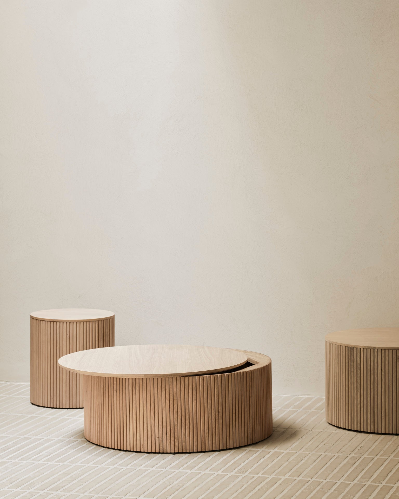 Three Ethnicraft Roller Max Coffee Tables in solid oak, each at a different height with vertical grooves, are displayed on a light patterned floor against a plain beige wall for a minimalist modern look.