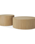 The Roller Max Coffee Tables by Ethnicraft are crafted from solid oak, featuring round shapes, vertical side grooves, and a smooth finish—one taller and smaller, the other wider and lower—for a modern minimalist look.