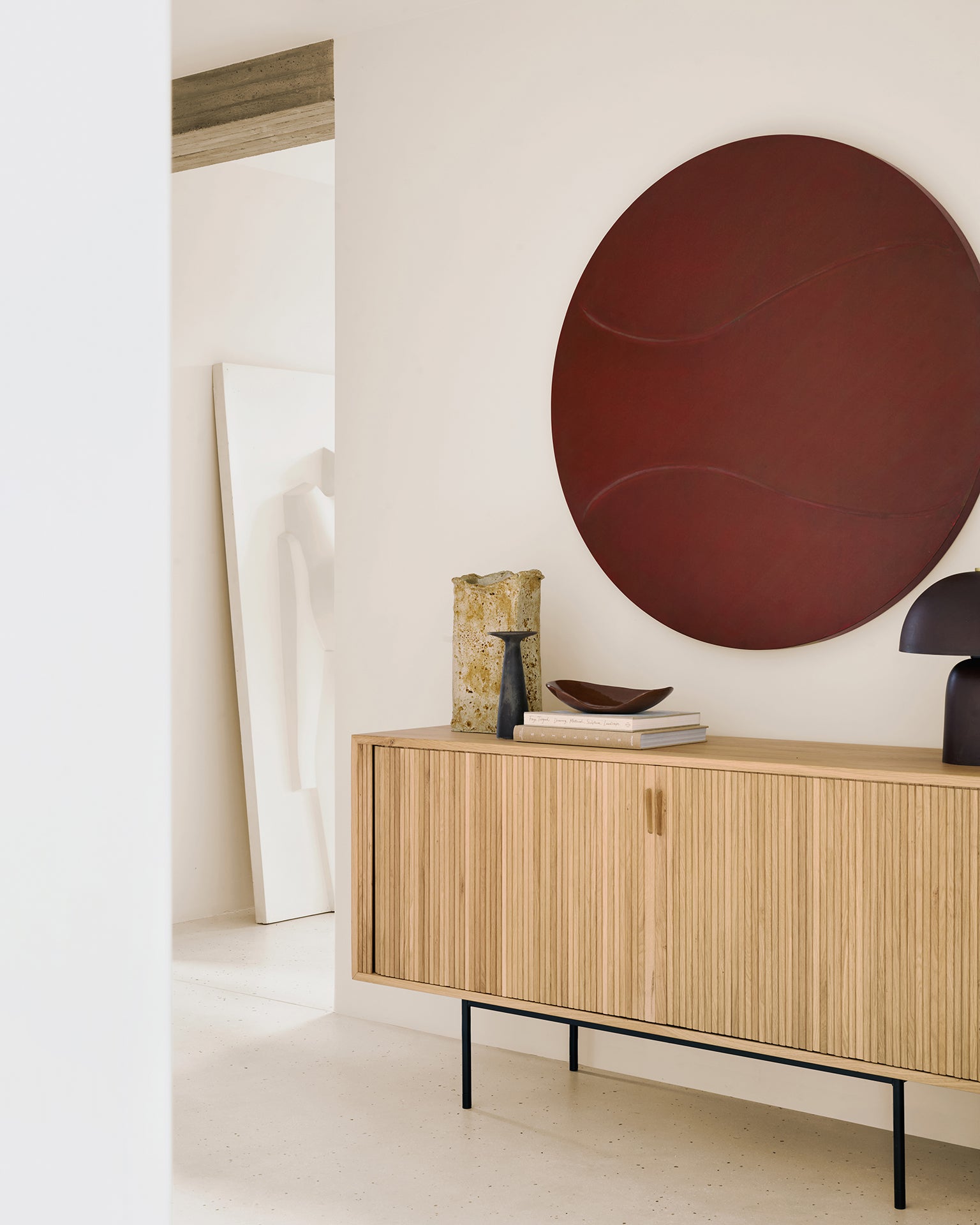 A minimalist interior features Ethnicraft's Roller Max Sideboard styled with stacked books, ceramics, a sculptural lamp, and bold round red wall art. Light walls and neutral tones amplify the airy ambiance.