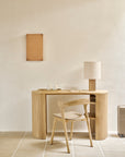 A minimalist workspace featuring the Roller Max Desk | Oak by Ethnicraft, paired with a matching chair, beige lamp, two cups, a notebook, and a suitcase placed on the right. A plain artwork hangs on the cream-colored wall, complementing the room’s calm and neutral color palette.