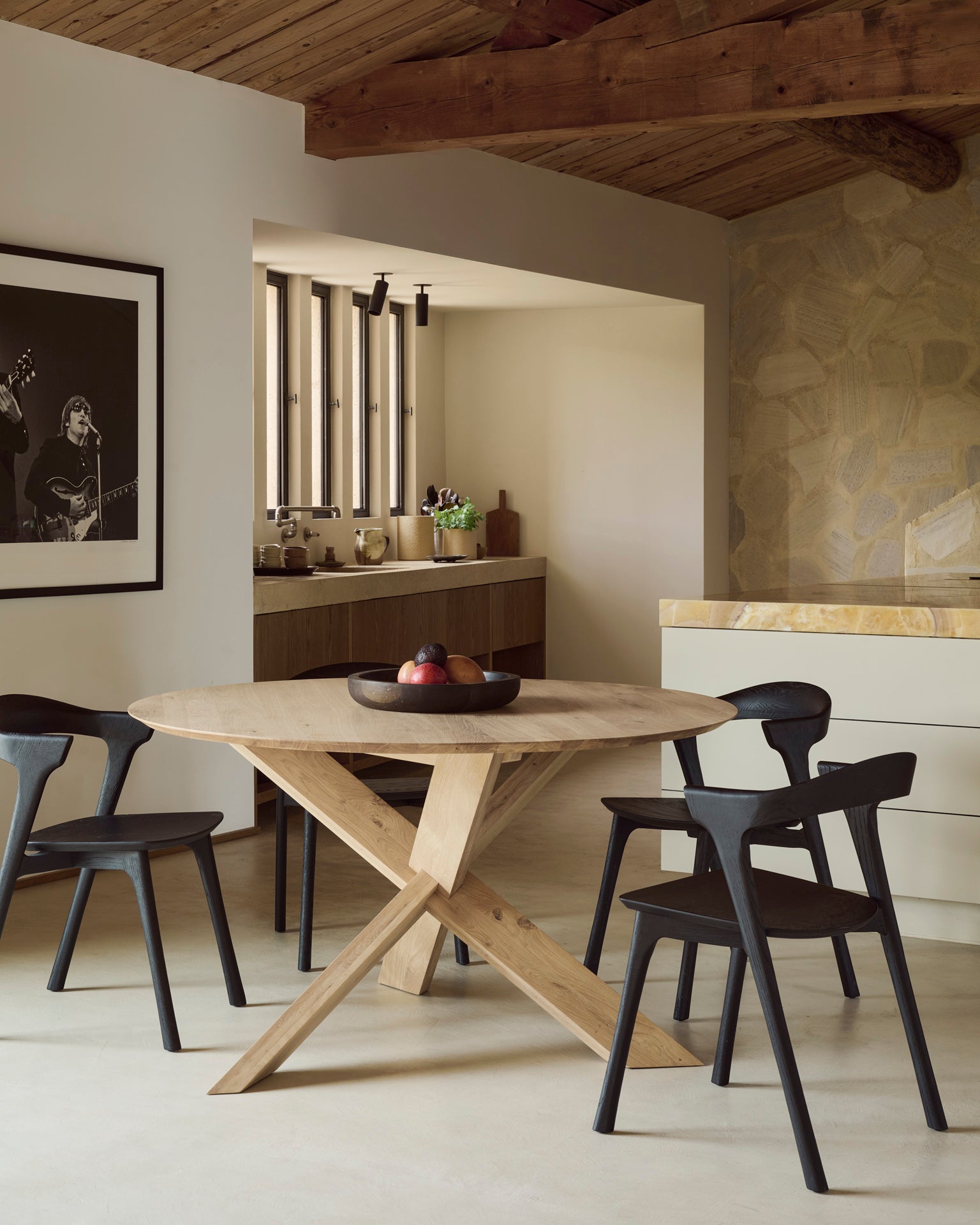 A modern dining area featuring the Ethnicraft Circle Dining Table in European oak, surrounded by four contract-grade black chairs and accented with a fruit bowl centerpiece. Natural wood and stone elements complement the space, while an Ethnicraft black-and-white photo adorns the wall near the open kitchen.