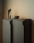 The Ethnicraft Stairs cupboard in Oak Brown stands against a light beige wall, topped with a round stone bowl and sculptural black decor, while soft natural light casts gentle shadows across its textured push-to-open doors.