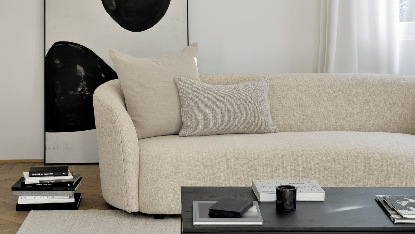 A minimalist living room with a light beige curved sofa, a low black coffee table, and stacked books is anchored by Ethnicraft’s Nomad Kilim Rug in Sand, while sheer white curtains let in natural light.