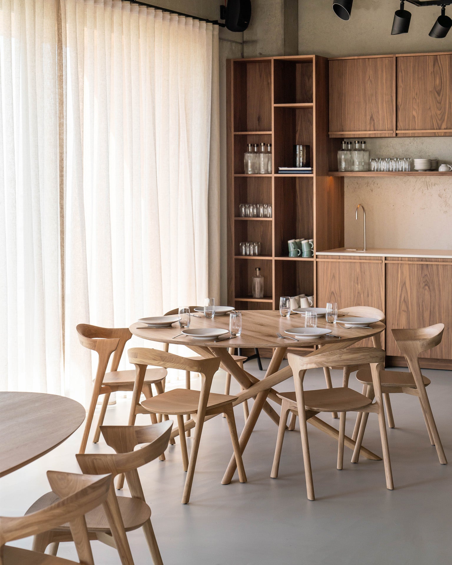A modern dining area features the Ethnicraft Mikado Dining Table Round in oak, four matching wooden chairs, tall shelves with glassware, and a wood-cabinet kitchenette. Large sheer curtains let natural light fill the minimalist space.