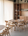A modern dining area features the Ethnicraft Mikado Dining Table Round in oak, four matching wooden chairs, tall shelves with glassware, and a wood-cabinet kitchenette. Large sheer curtains let natural light fill the minimalist space.