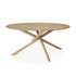 The Mikado Dining Table Round - Oak by Ethnicraft features a solid oak build with a smooth top and three angled legs meeting at the center, embodying modern minimalist design.
