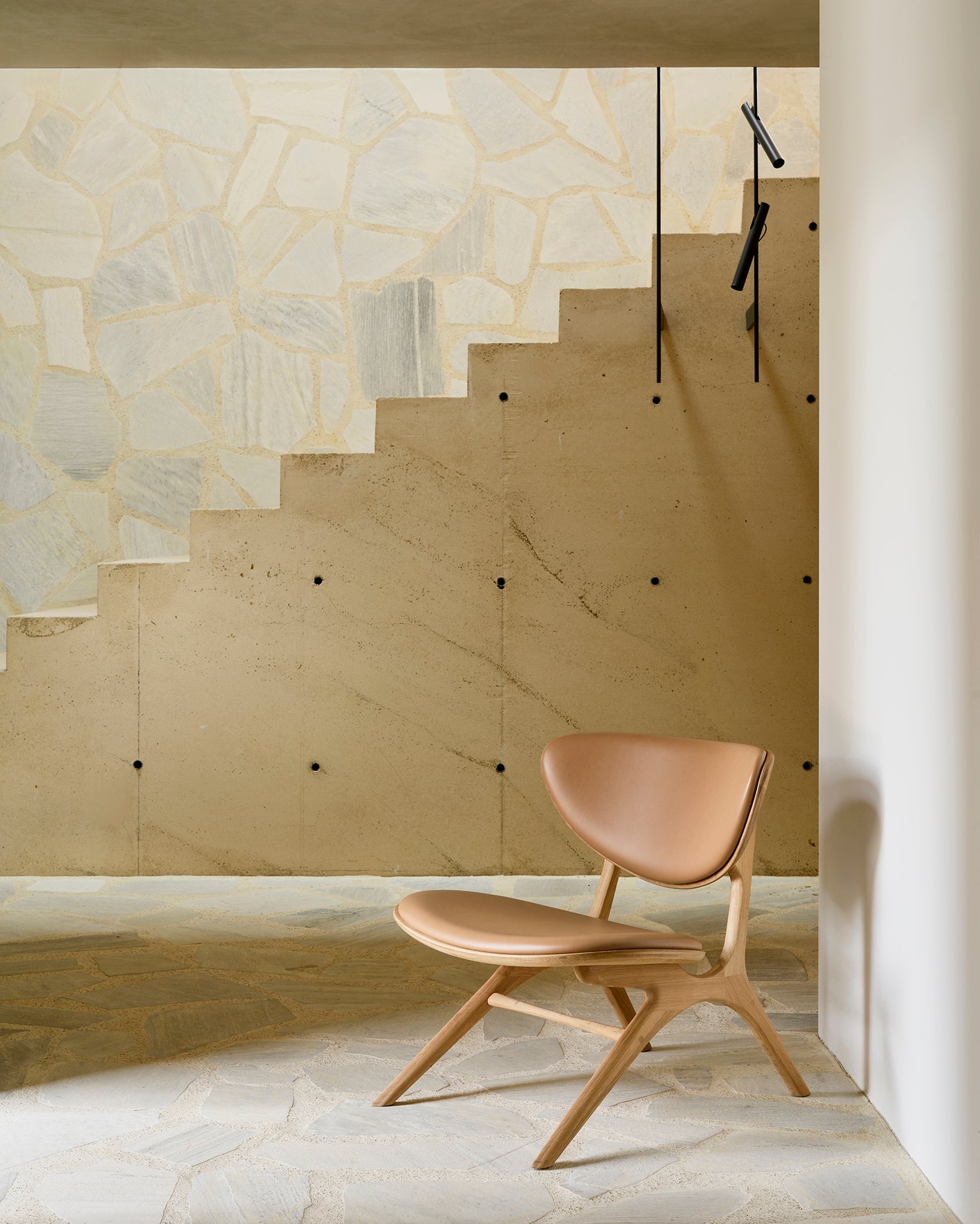 The Ethnicraft Eye Lounge Chair - Oak with Leather in tan stands beside a beige stone staircase with geometric light-colored stone wall accents and a minimalist black railing in a bright, modern interior.