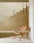 The Ethnicraft Eye Lounge Chair - Oak with Leather in tan stands beside a beige stone staircase with geometric light-colored stone wall accents and a minimalist black railing in a bright, modern interior.