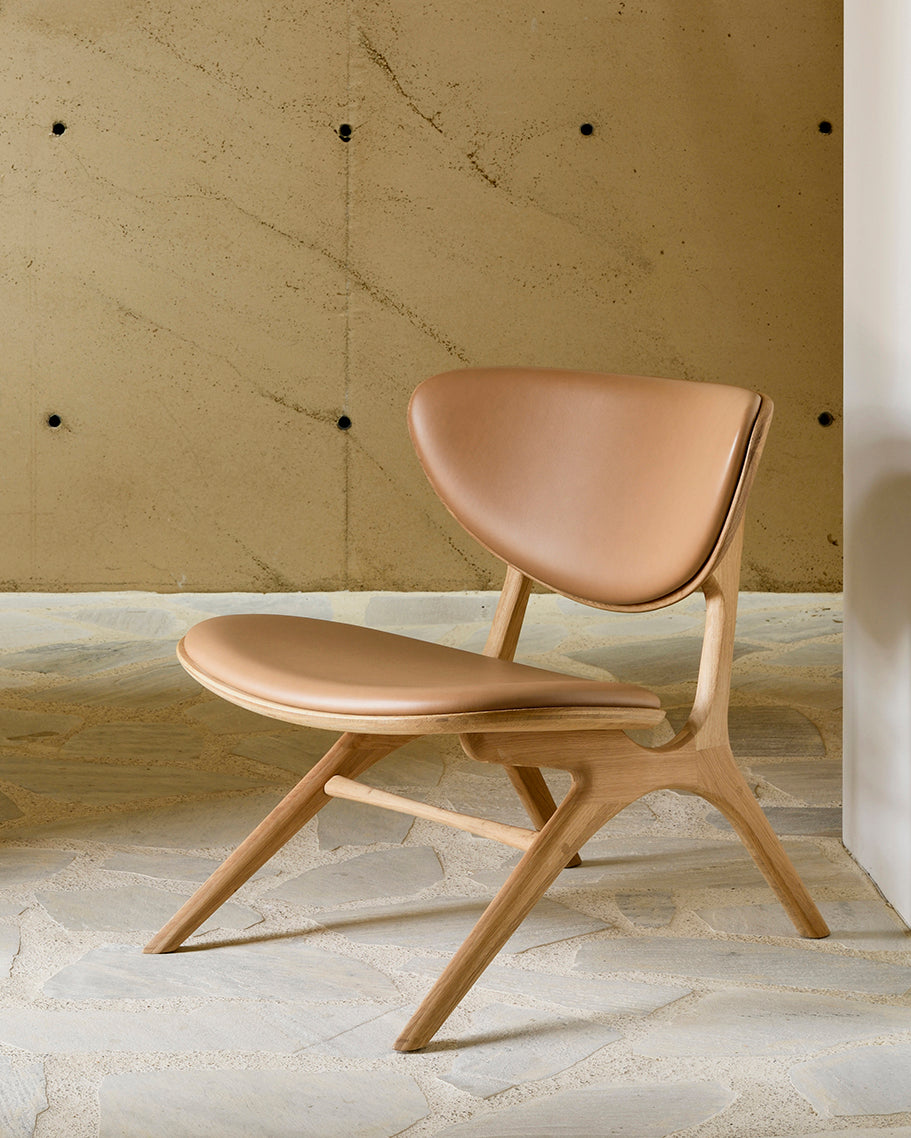 The Ethnicraft Eye Lounge Chair - Oak with Leather sits on a stone floor against a beige wall with visible dark bolts.
