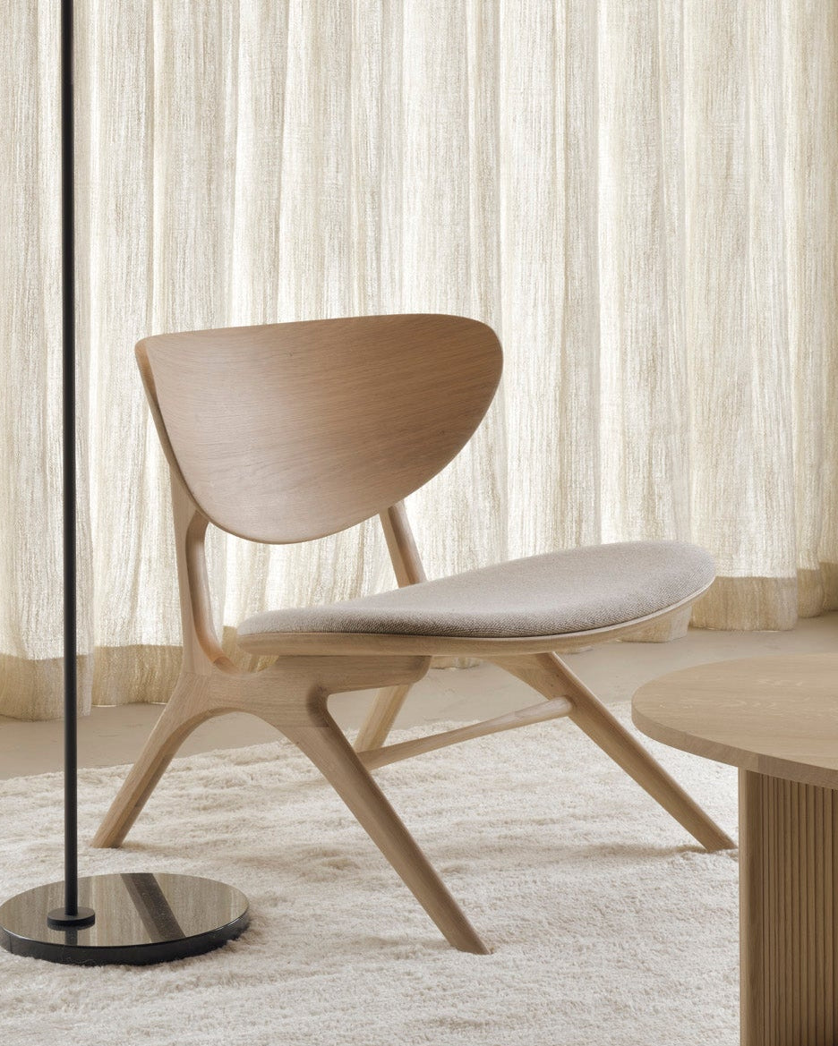 A minimalist living space featuring the Ethnicraft Eye Lounge Chair in oak with off-white fabric, a black floor lamp, a round wooden coffee table, and beige curtains. The room showcases neutral tones and modern, Scandinavian-style decor.