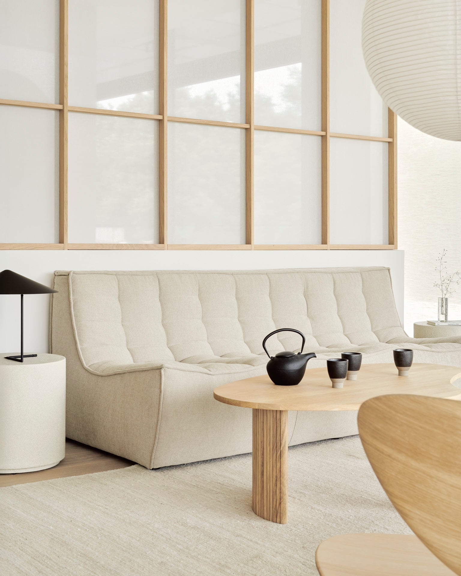 A minimalist living room features the Ethnicraft N701 Modular Sofa in beige, a round wooden coffee table with a black teapot and cups, a modern black lamp, and wooden-framed glass panels—all in neutral European-inspired tones.
