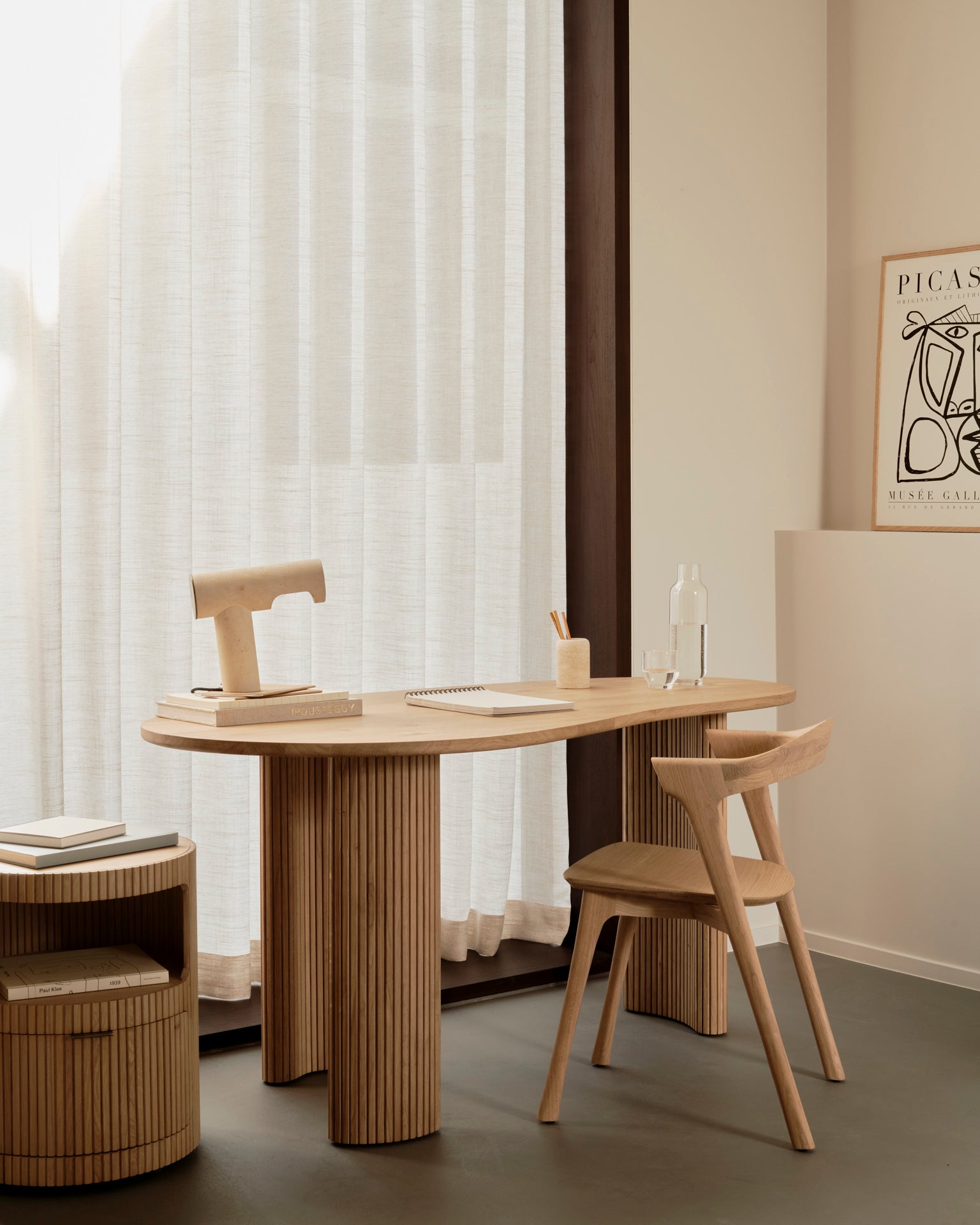 Minimalist home office with an Ethnicraft light wood desk and chair, Ethnicraft Boomerang Drawer Unit – Oak, notepad, book, sculptural lamp, water bottle, sheer curtains filtering light, and a Picasso print on the beige wall.