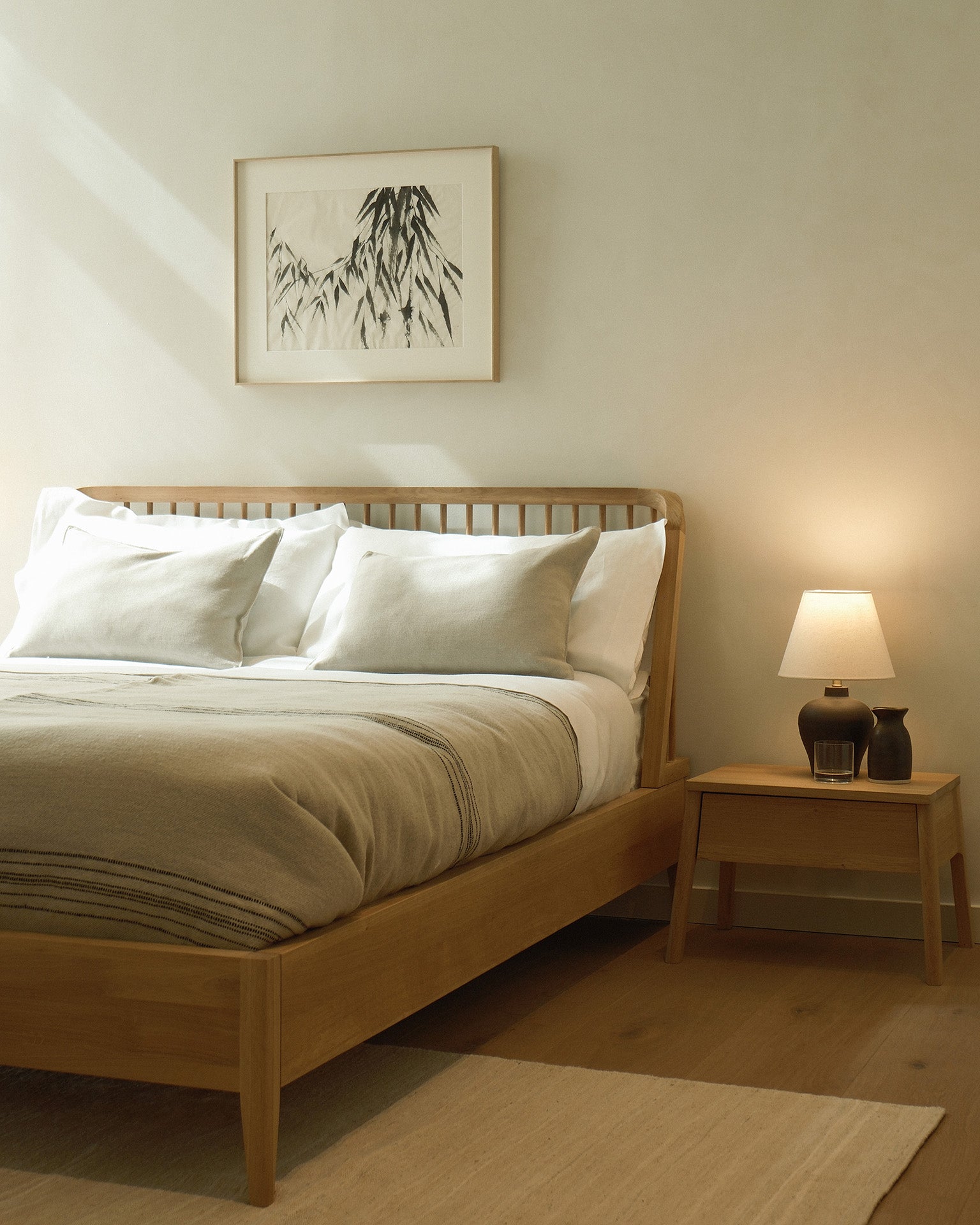 A minimalist bedroom features the Ethnicraft Spindle Bed - Oak, white and beige bedding, a lamp-topped bedside table, and framed abstract art on a pale wall. Soft natural light gently brightens the space.