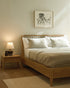 A minimalist bedroom features the Ethnicraft Spindle Bed - Oak, white and beige bedding, a lamp-topped bedside table, and framed abstract art on a pale wall. Soft natural light gently brightens the space.