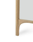 A close-up of the Ethnicraft PI Floor Mirror - Oak captures the smooth, light oak wood with curved detailing, a polished yet imperfect finish at the bottom corner, and part of the reflective glass.