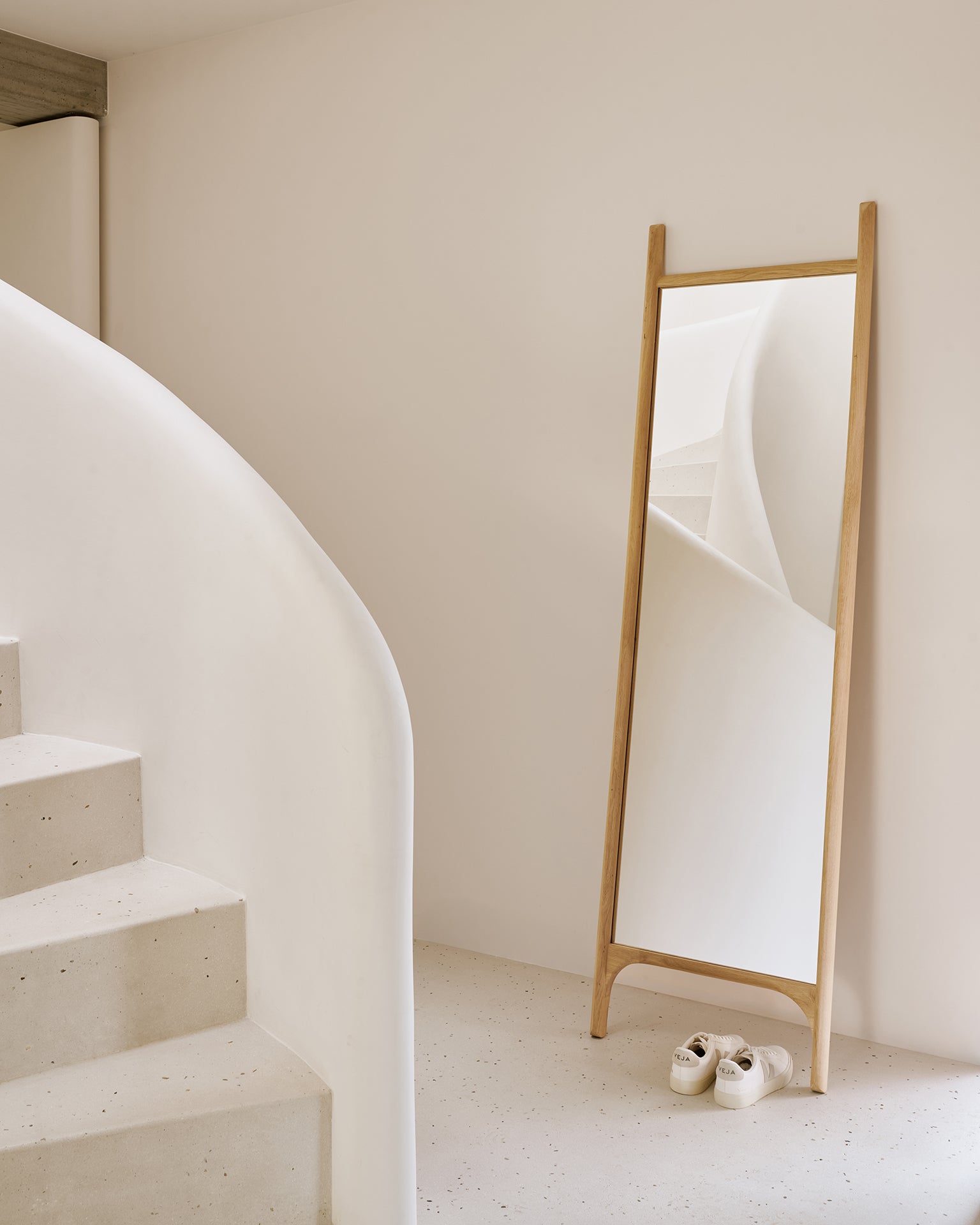 A minimalist interior features a curved white staircase, the Ethnicraft PI Floor Mirror in oak reflecting the stairs, and a pair of white sneakers neatly placed on the polished imperfect floor before the mirror.